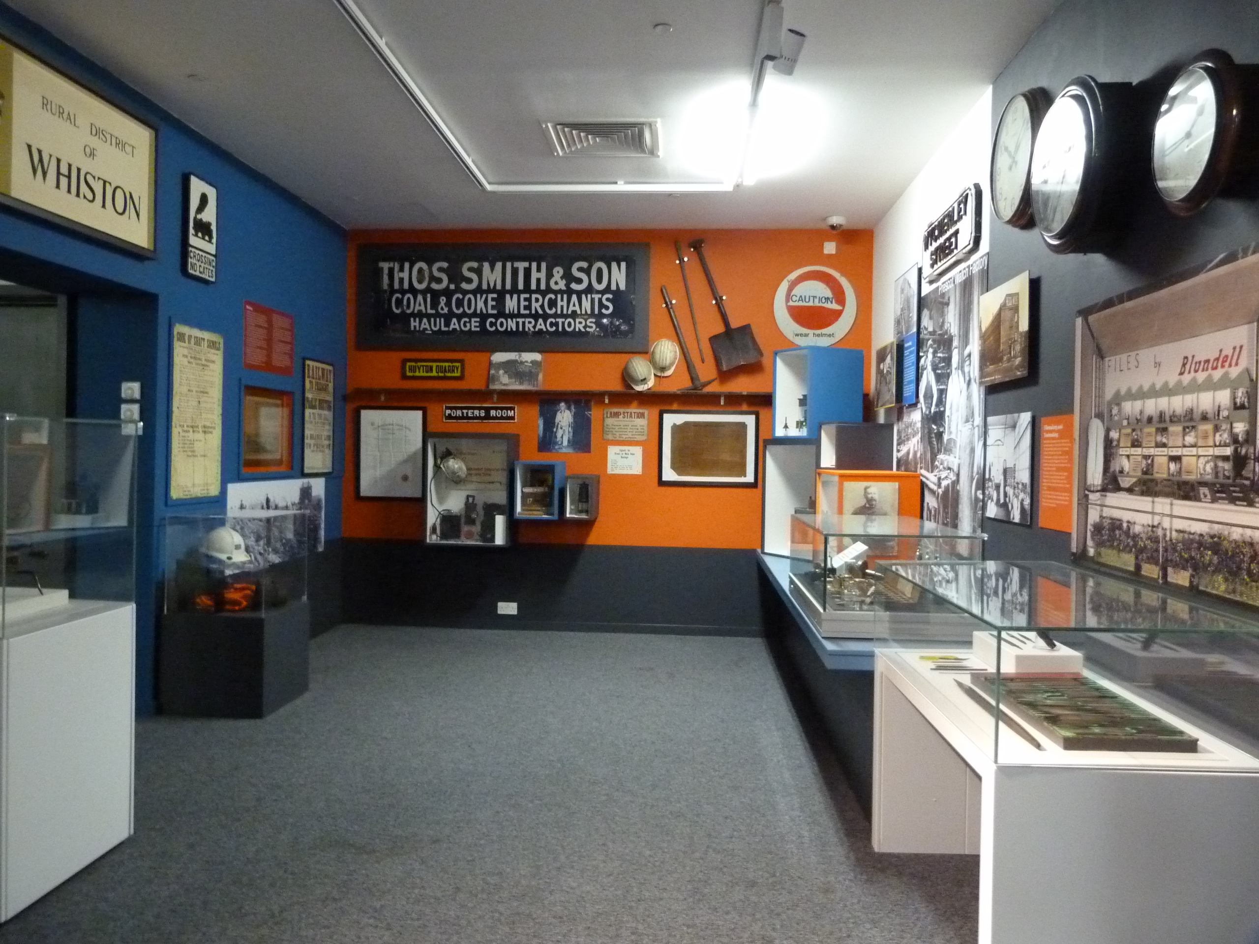 Photograph of display cases and wall-mounted exhibits at Prescot Museum, showing coal excavation tools and safety wear.