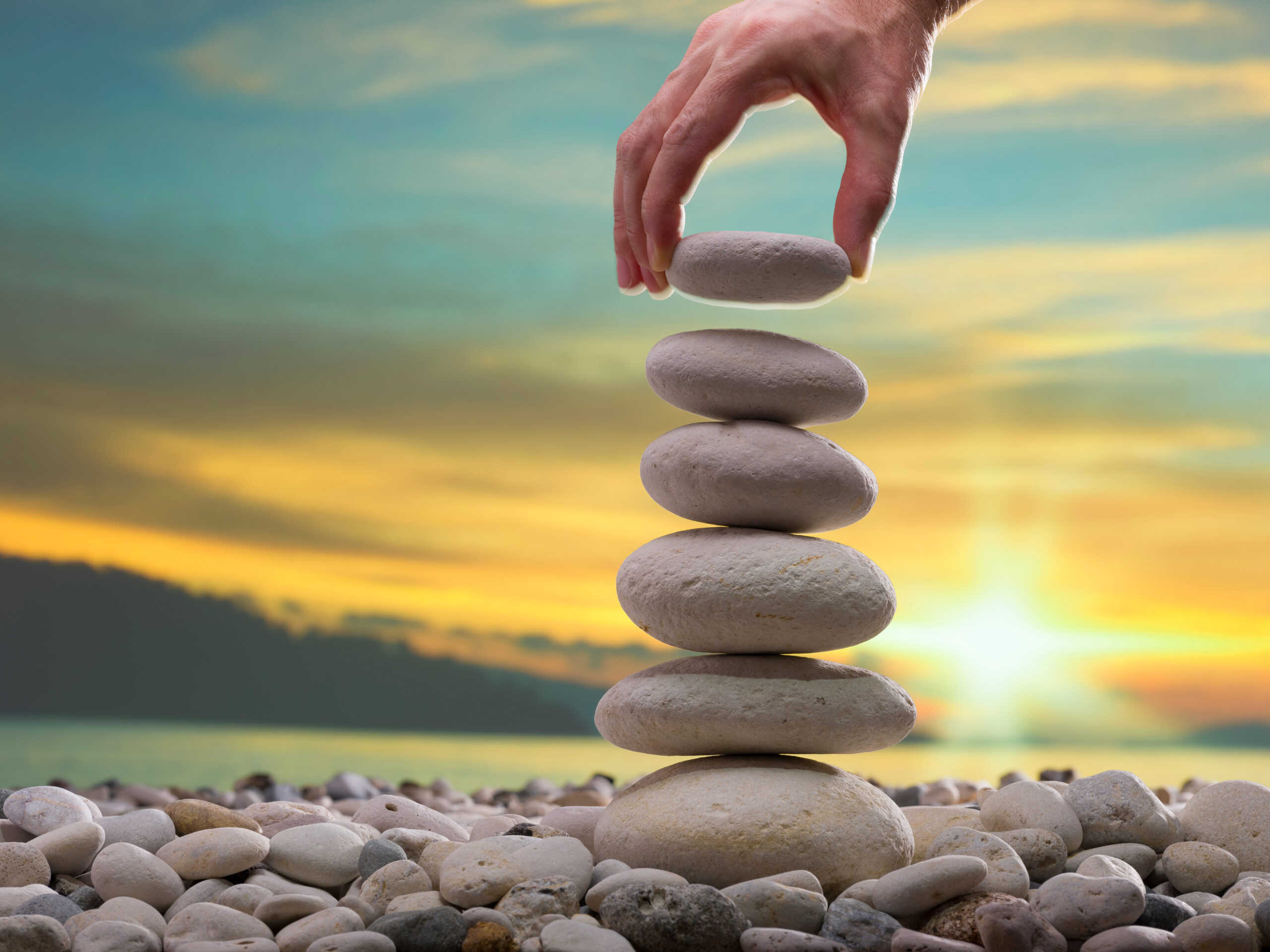 A photograph of a persons hand making a tower of pebbles outdoors, placing the last pebble on top of the tower.