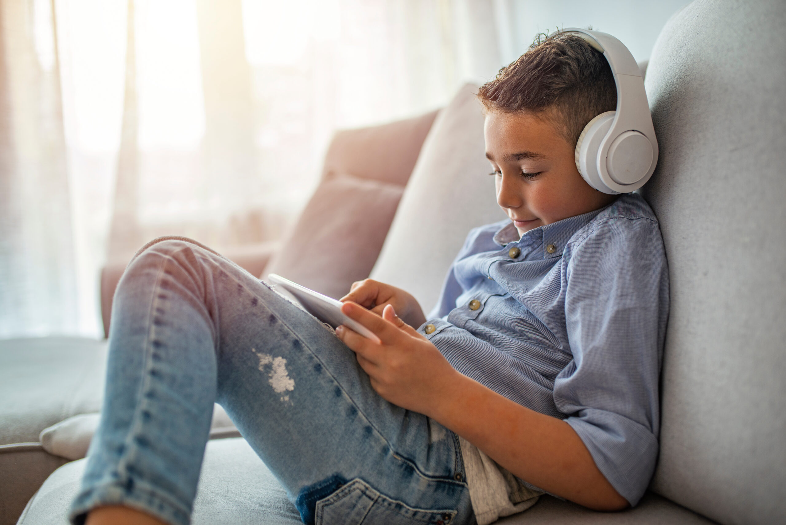 Little boy aged 6 listening to the music using modern tablet. The boy is wearing headphones and looking at tablet. Young boy with headphones and tablet listening to audiobook at home