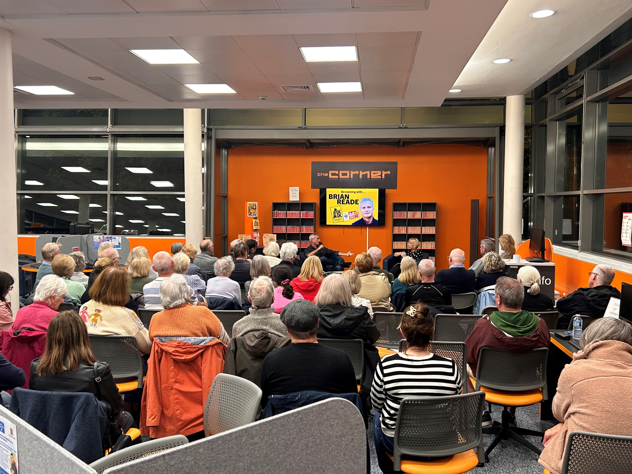 A photograph from the point of view at the back row of the audience listening to Brian Reade sitting and talking into a microphone.